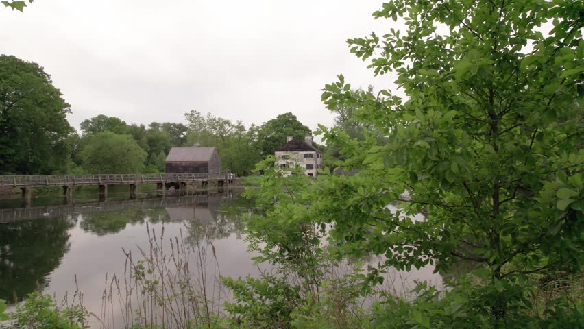 A serene landscape view of the historical Philipsburg Manor in Tarrytown, NY. The scene captures a calm pond reflecting a wooden mill, white manor house, and a timber bridge, surrounded by dense green trees and foliage on a cloudy, overcast day.