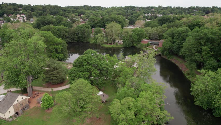 An aerial drone shot glides over the lush greenery and calm waters of the Philipsburg Manor historic site in Sleepy Hollow, New York. The footage features the stone manor house, a wooden bridge across the mill pond, and the historic gristmill surrounded by a dense forest landscape.