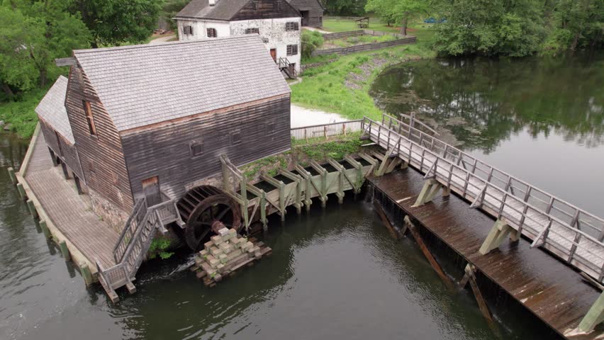An aerial drone shot reveals the historic Philipsburg Manor grist mill and its large wooden waterwheel. The scene features a weathered wooden bridge and a calm river surrounded by lush summer greenery, showcasing colonial American architecture and 18th-century industrial technology.
