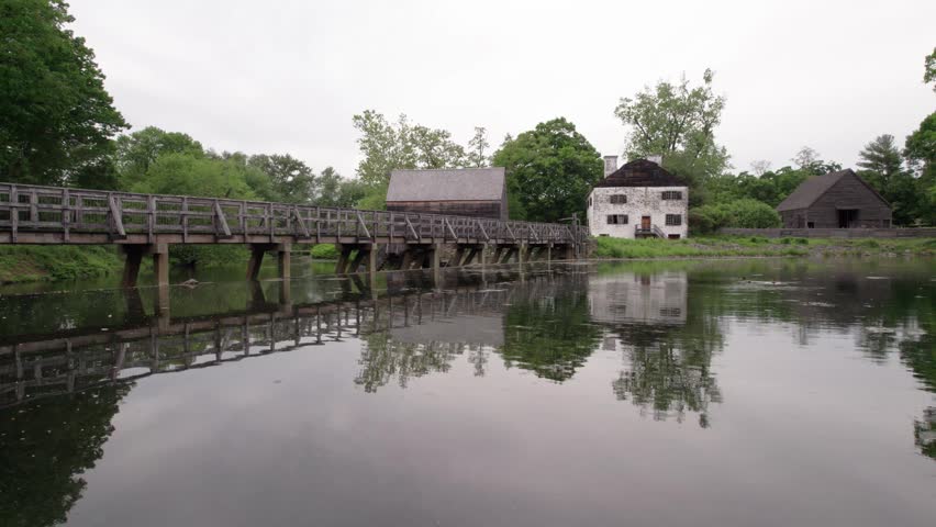 A serene view of the historic Philipsburg Manor in Sleepy Hollow, featuring a colonial-era manor house, a wooden bridge, and traditional outbuildings. The calm millpond perfectly reflects the architecture and lush green surroundings under an overcast sky during a slow panning shot.