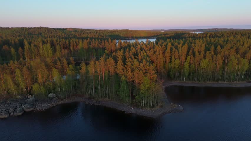 Drone reverse rise over Puumala, Lake Saimaa, Finland, revealing pine forest islands, rocky shoreline, and wide lake waters in warm evening light.