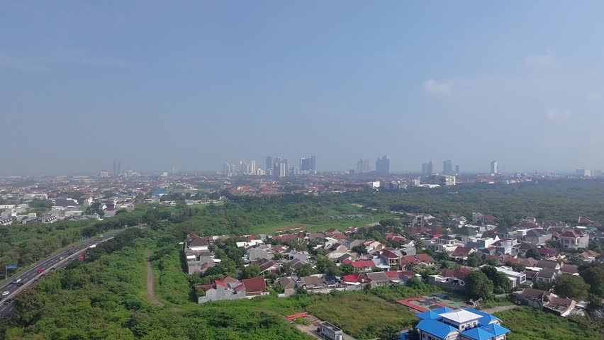 Aerial view of a city showing residential neighborhoods with houses and red-tiled roofs, surrounded by dense green trees and grassy areas. A multi-lane highway with vehicle traffic runs through the greenbelt. In the far background, a skyline of multiple high-rise buildings is visible under a clear blue sky.