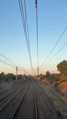 Straight railway tracks disappearing into the horizon at sunset with overhead electric power lines and warm evening light. Scenic railroad perspective through rural landscape featuring transportation infrastructure, symmetry and travel concept in vertical composition.