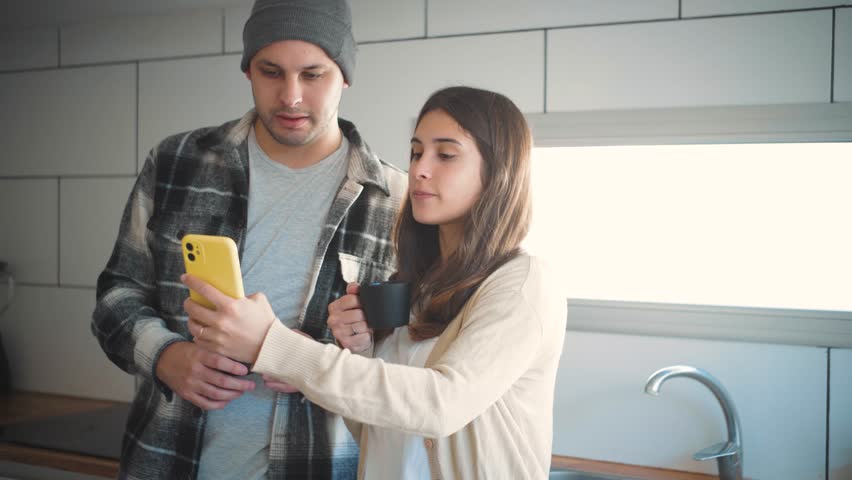 4K video of a young Hispanic couple drinking coffee while laughing and watching social media reels on their mobile phone. Concept of everyday lifestyle, emotional wellbeing, and a healthy romantic relationship.

