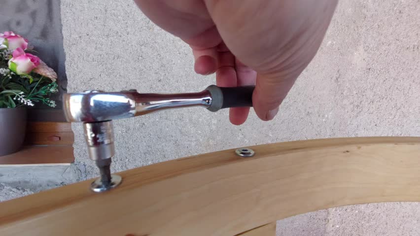 Man assembling wooden furniture frame with a socket wrench ratchet outdoors.