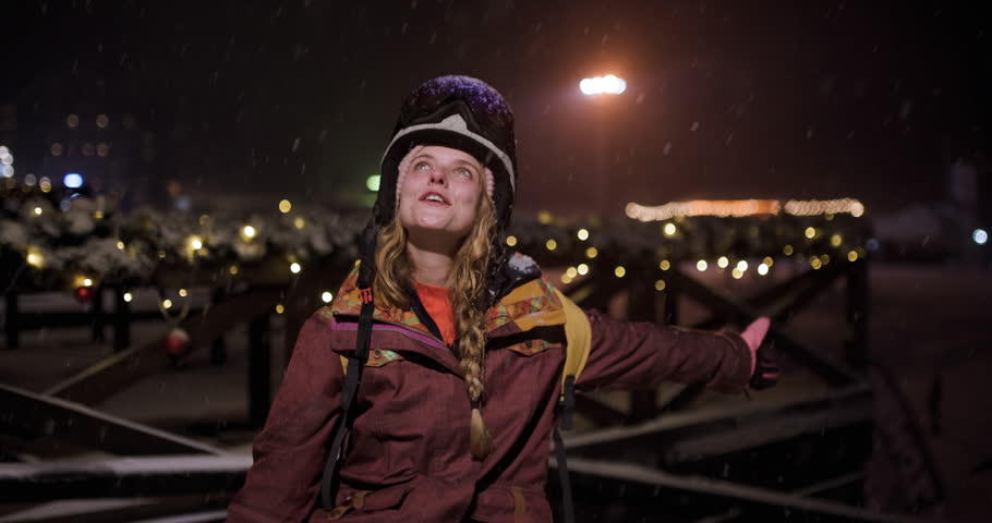 Close-up portrait of a happy young woman snowboarder looking up at the falling snow and smiling at the camera at night in a ski resort.