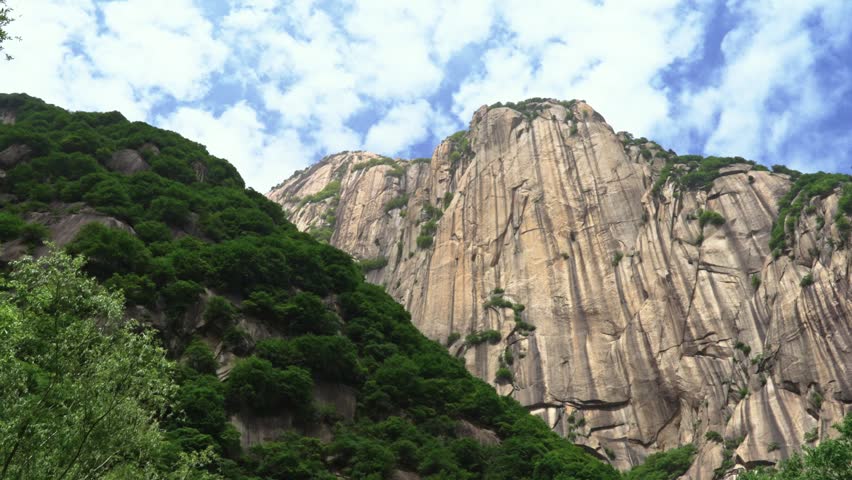A crisp low-angle panning shot looking up at a massive, vertical granite rock face on Mount Hua under a bright blue sky filled with moving white puffy clouds.