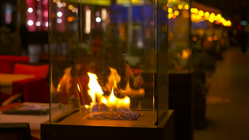 Decorative glass tabletop fireplace burning at night. Warm fire burning in a modern glass and metal decorative bio fireplace on a restaurant table, with a blurred city street
