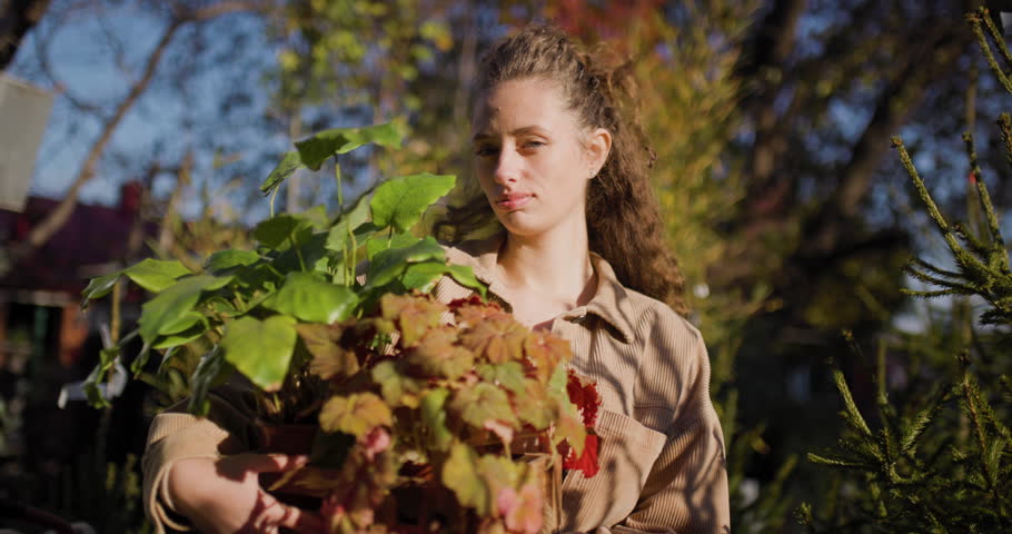 A young woman smiles while holding potted plants in a garden center, surrounded by lush greenery and vibrant foliage, capturing a joyful connection with nature.