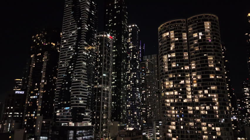 Ascending aerial view of illuminated residential skyscrapers and modern city skyline at night in Melbourne, Australia.