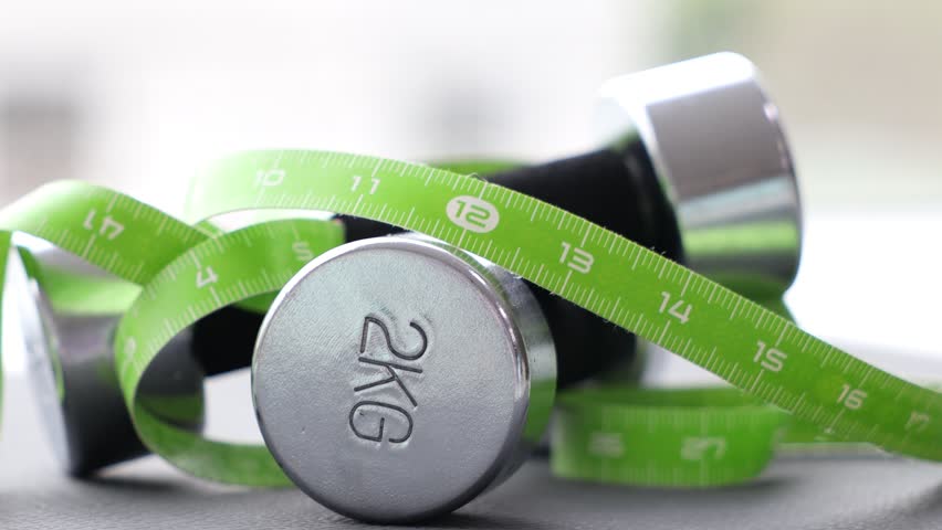 Measuring tape and dumbbells on blurred background, closeup. Camera moving left