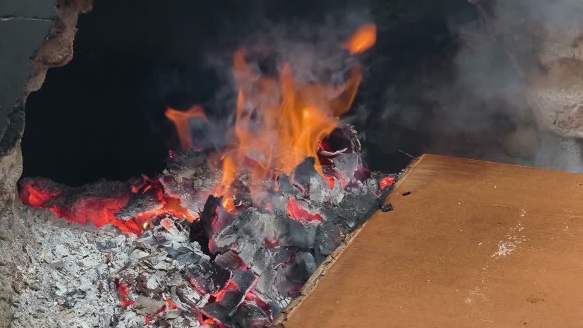 A close-up view of orange flames flickering amidst glowing embers and wisps of grey smoke inside a stone furnace. A piece of wood sits at the edge of the fire, showing the intense heat and combustion process in a dark environment.