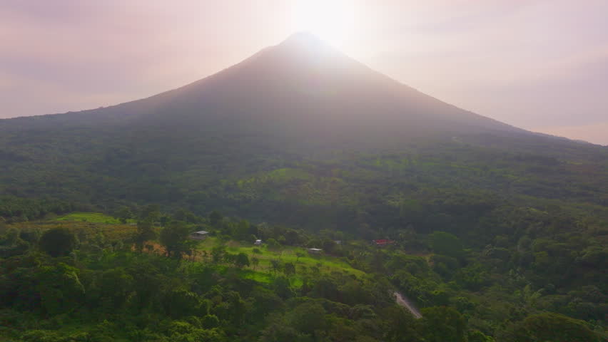 Aerial drone footage captures the majestic Arenal Volcano at sunrise in Costa Rica. Sun rays illuminate the lush green rainforest and small rural houses nestled in the tropical landscape below.