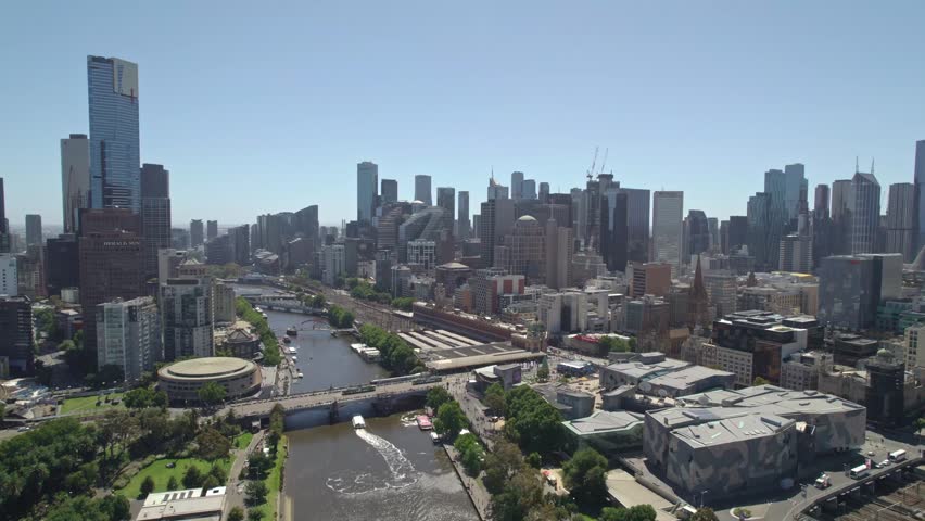 Cinematic aerial shot capturing metropolitan landscape of Melbourne city. Wide panorama shows skyscrapers, tranquil Yarra river, busy bridges, and green urban parks under bright summer sun.