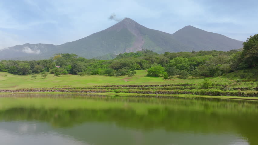 Aerial drone footage panning across a scenic golf course with calm reflective pond and stone terraces, revealing majestic smoking volcano peaks rising above dense tropical rainforest under hazy skies.