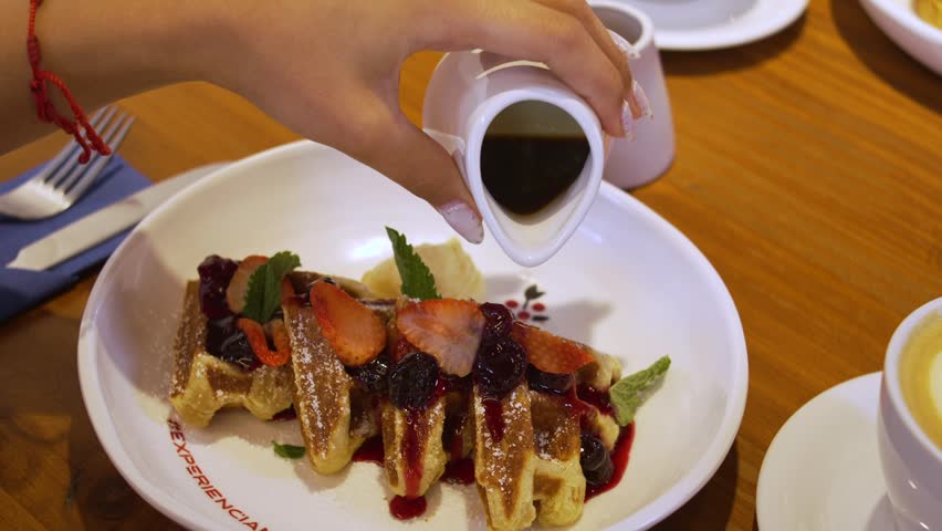 Person Drizzling Dark Syrup on Belgian Waffles Topped with Fresh Fruit at Restaurant Brunch Table