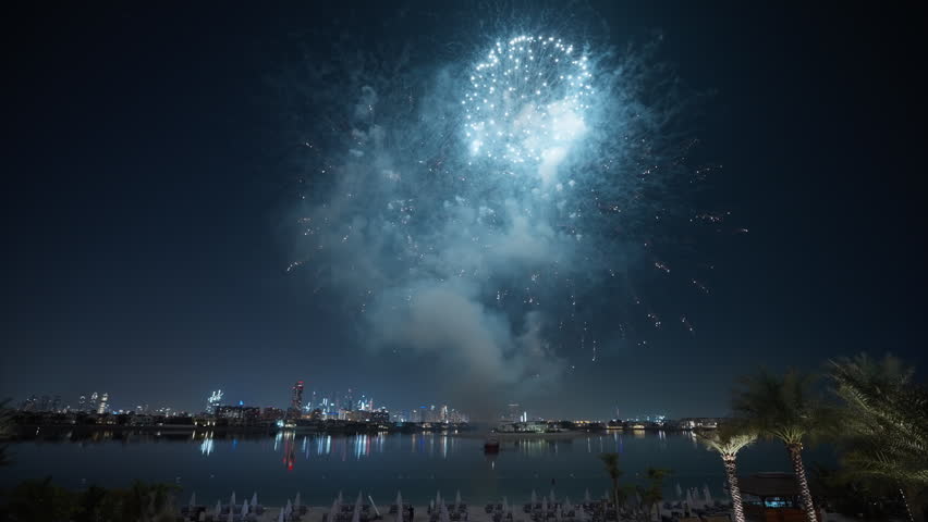 Vibrant fireworks explode in brilliant pink red and white bursts over Dubai waterfront. City skyline reflects on calm water as palm trees frame the stunning pyrotechnic display against dark night sky.