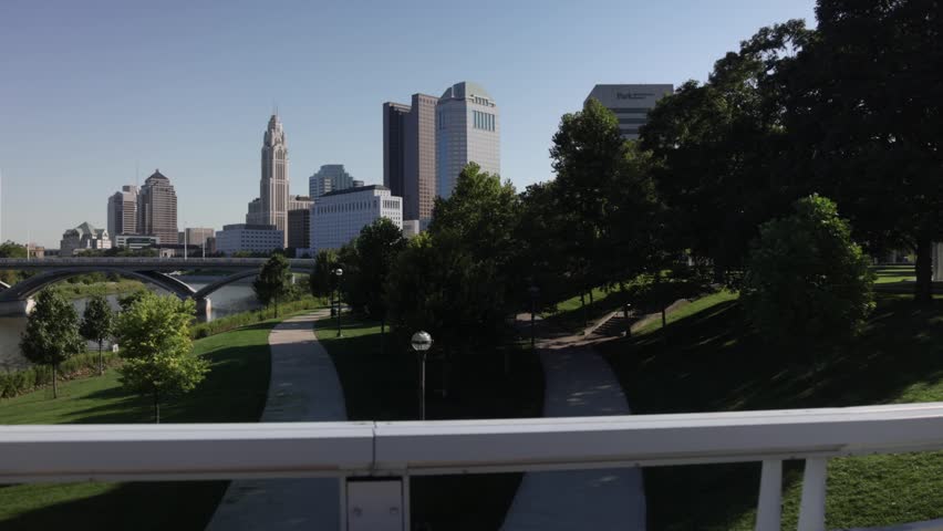 Dynamic outdoor video clip offering panoramic view of city skyline with tall buildings, a river with a bridge, and lush green parkland with walking paths on a sunny day.