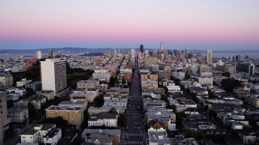 Aerial drone footage sweeps over San Francisco cityscape. Dusk sky with purple and pink hues. City buildings and hills visible. Transportation on streets.