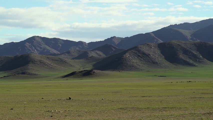 Panoramic Mongolia steppe shows rolling mountains and vast green plains in empty countryside. Wide grassland landscape stretches across untouched wilderness with natural scale. The broad composition highlights uninterrupted grassland, rolling slopes, and distant highlands across open Mongolia. Search relevance includes steppe biome, pastoral meadow, Asian prairie, expansive scenery, wild terrain, and natural horizon. The clip can function as an establishing shot for travel or documentary projects.