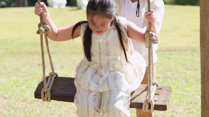 A young boy pushes his smiling sister on a wooden swing in a bright park