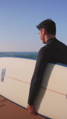 vertical Young man in a wetsuit holding his surfboard while standing barefoot on the sand at the beach, looking over his shoulder at the camera with the ocean and waves in the background at sunset