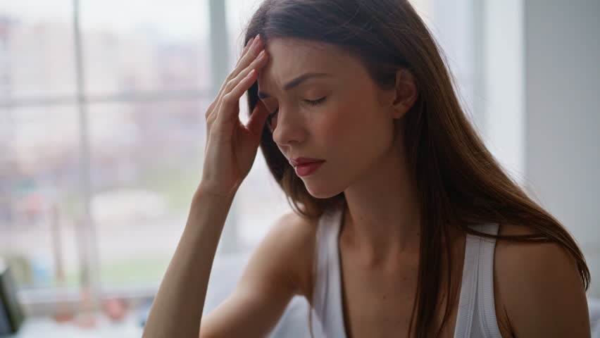 Awake woman massaging temples at wakeup on bed closeup. Stressed lady suffering of migraine in light bedroom sitting on comfortable bedding. Tired brunette feeling headache touching head in bedroom