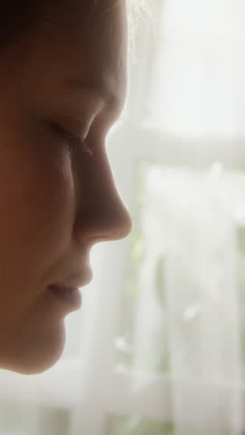 Vertical side view closeup shot of Caucasian girl smelling white aster flower blissfully against window at home, scene lit with warm hazy lighting, story format