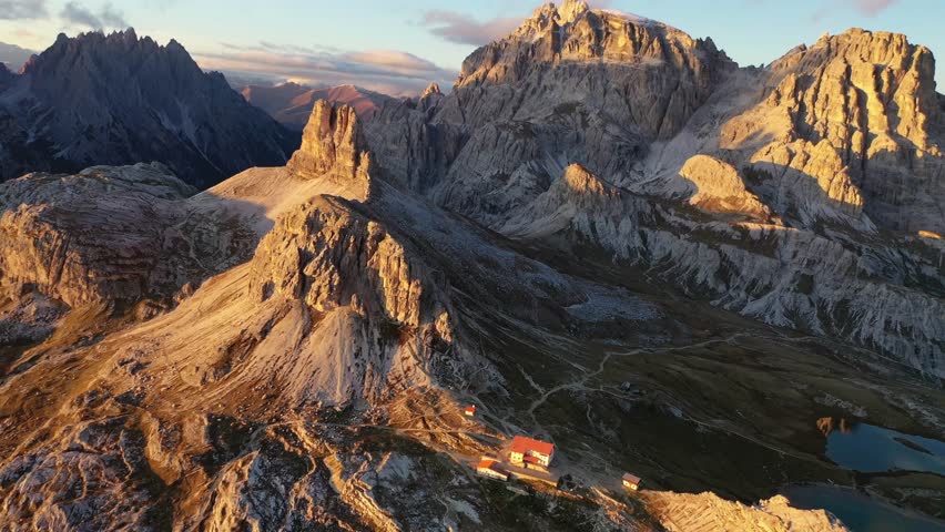 Breathtaking high-angle drone shot of the remote Rifugio Locatelli alpine hut and Monte Paterno peak in Italy. Features rugged limestone rocks illuminated by warm golden hour sunset sunlight.