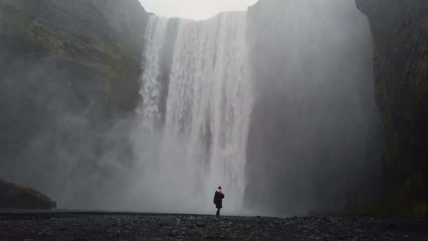 Lone woman standing in front of the huge Skogafoss waterfall in Iceland. The tourist is raising her arms in joy and freedom