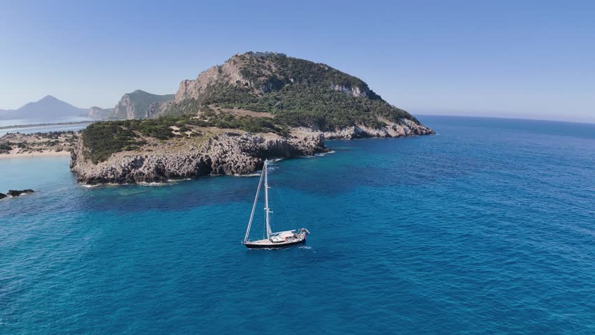 Beautiful cinematic drone footage captured above Voidokilia Beach and the surrounding Mediterranean coastline in Messinia, Peloponnese, Greece. The video features a luxury sailing yacht navigating deep blue Ionian Sea water beside rugged rocky cliffs and turquoise coastal shallows. In the background, the iconic curved shoreline of Voidokilia Bay, sandy dunes, Gialova Lagoon and distant mountain ranges create a dramatic and atmospheric natural panorama. Perfect for travel films, luxury tourism advertising, sailing lifestyle content, yacht promotions, Mediterranean vacation visuals and cinematic aerial establishing shots.