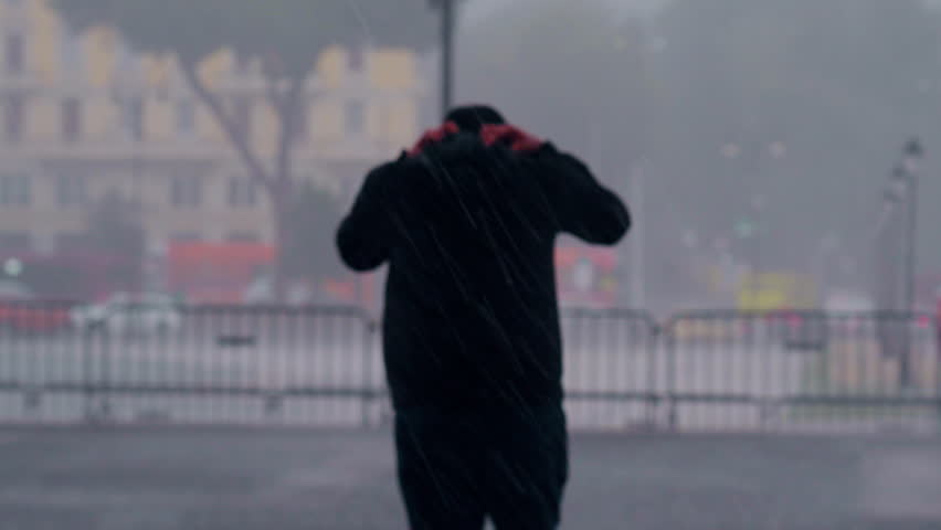 Person walking away on wet cobblestone street during heavy rain, holding hood tightly for protection, with blurred city traffic and barriers in the stormy background