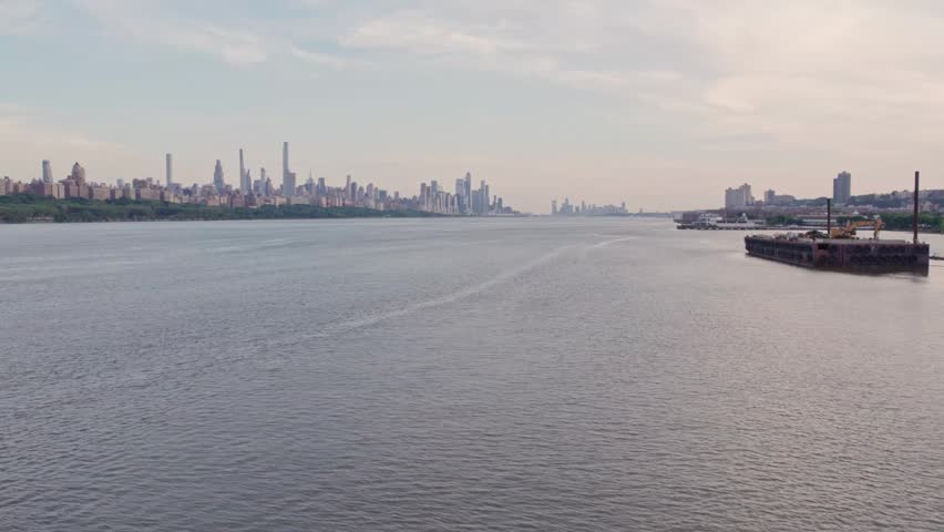 A wide aerial drone shot captures the iconic New York City skyline across the expansive Hudson River. The scene features the dense skyscrapers of Manhattan under a soft, overcast sky, with the calm river water reflecting the ambient light and a glimpse of the New Jersey shoreline.