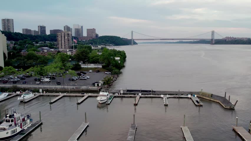 This aerial drone footage captures a peaceful marina on the Hudson River, featuring several boats docked along the pier. In the background, the iconic George Washington Bridge spans the river under a soft, overcast sky, connecting the New Jersey coastline to New York City.