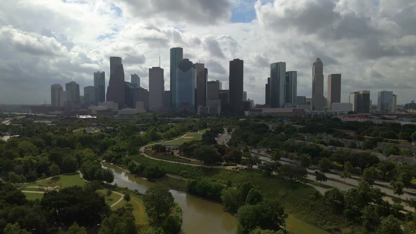 Dramatic aerial shot of Houston cityscape featuring prominent skyscrapers under cloudy sky. Dynamic view captures lush greenery of Buffalo Bayou Park and winding river in foreground of city.