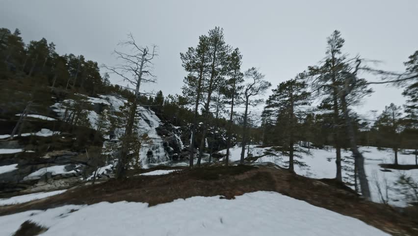 Smooth aerial FPV alongside a rushing winter waterfall, tracking fast-flowing rapids through a serene, snowy wilderness