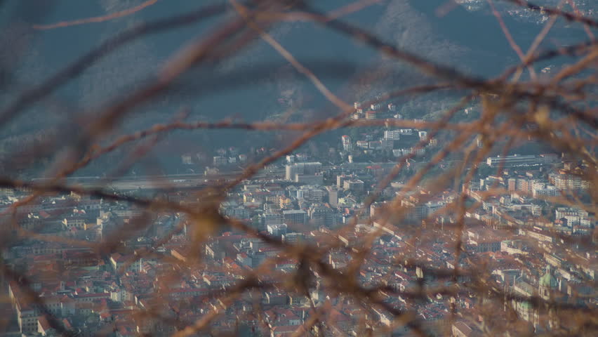 Lake town close up seen through plant branches foreground, handheld panning shot. Detail of lakeside mountain city skyline framed by green leaves.