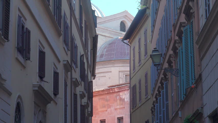 Narrow alleyway between tall buildings leading to dome of Renaissance church in Rome, Italy, with warm pastel walls, shutters, and nostalgic Mediterranean atmosphere