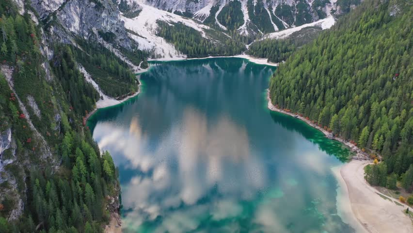 Stunning high-angle aerial drone shot of the emerald Lago di Braies lake in the Italian Dolomites. Features a thick green pine forest lining the water with the massive Croda del Becco peak above.