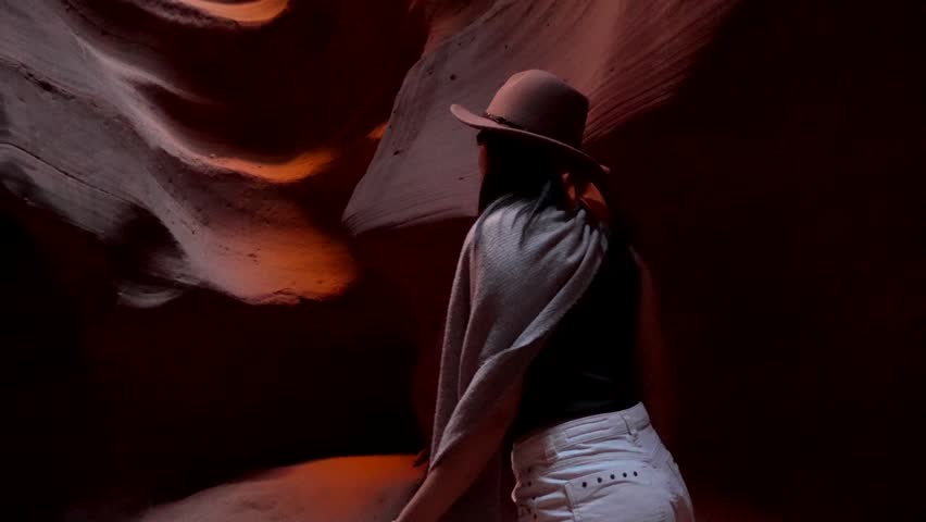 Beautiful tourist woman with a hat walking inside the amazing red rock walls of upper antelope canyon, looking up at the sunlight filtering through the narrow passage in page, arizona