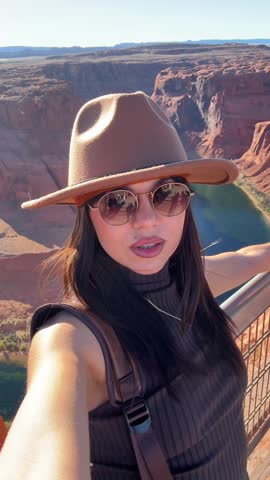Stylish young woman wearing a hat and sunglasses taking a selfie video with the beautiful landscape of horseshoe bend and the colorado river in page, arizona, creating memories of her vacation