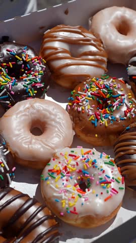 Top down view of a hand taking a chocolate glazed donut with rainbow sprinkles from an assorted box of delicious pastries, revealing the variety of sweet, sugary desserts available