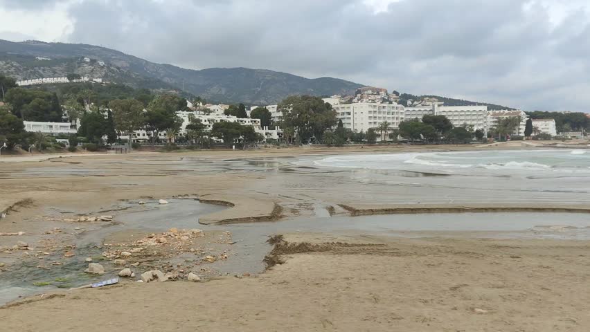 Freshwater stream eroding the sand and flowing across a beach into the ocean on a cloudy day, creating channels and rivulets with a coastal town and hills visible in the background