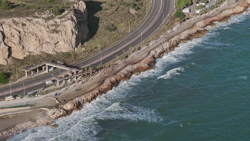 Aerial view of coastal highway and rocky shoreline in Malaga, Spain, with turquoise sea waves and dramatic Mediterranean seaside landscape.