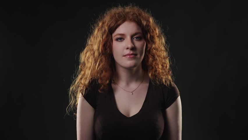 Emotional expression and human connection are conveyed through this studio portrait of a young woman with vibrant red curly hair. She smiles genuinely while looking directly at the camera against a st