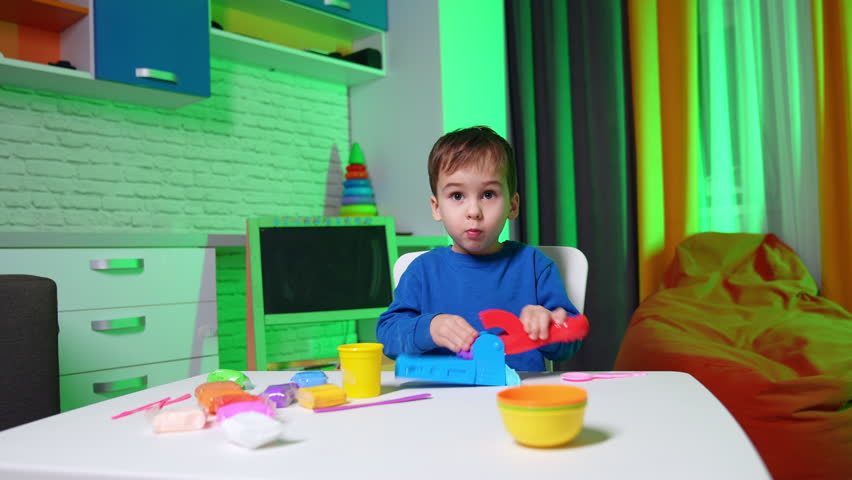 Adorable smiling Caucasian toddler in blue sweater sits at desk. Cute child uses molding machine to make plasticine figures.