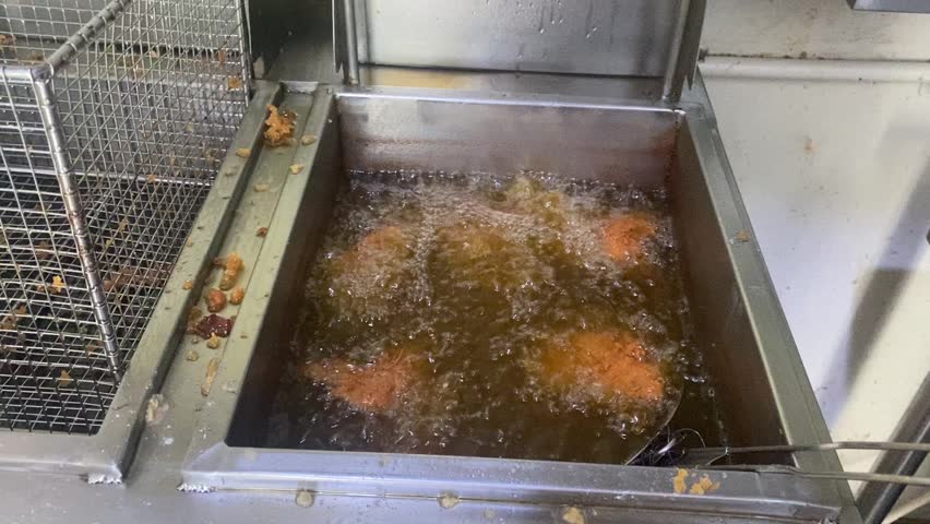 A person using a metal strainer to flip and turn pieces of chicken frying inside a commercial deep fryer with hot boiling oil.