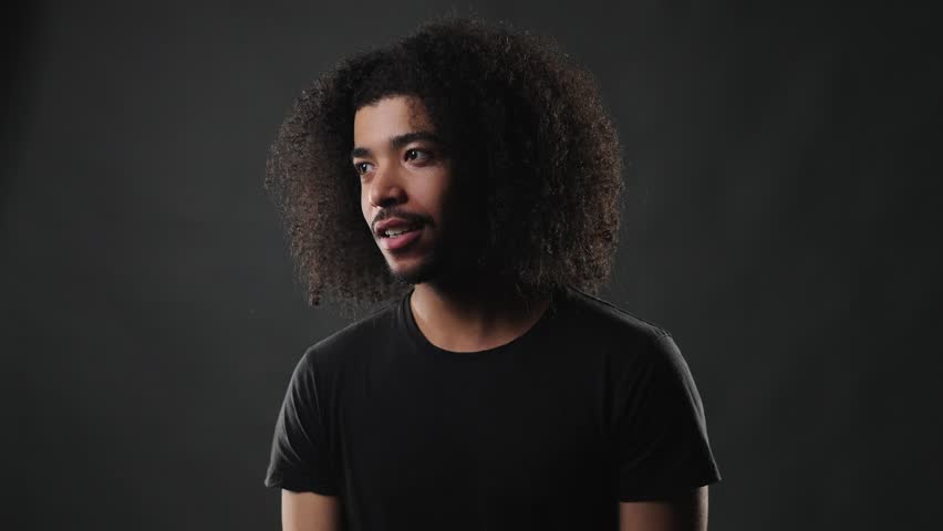 A thoughtful young African American man with voluminous curly hair and a simple black t shirt is captured in a studio portrait. He appears to be pondering deeply, with a serious and contemplative expr