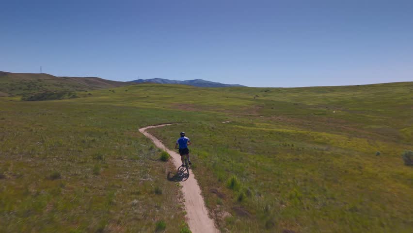 Dynamic drone chase shot following a mountain biker skillfully navigating twisting, winding singletrack trails in the Military Reserve, Boise, Idaho. The rider flows through the technical turns and rollers while downtown Boise skyline stands visible in the distance. Energetic spring action scene perfect for mountain biking, adventure sports, outdoor fitness, and trail lifestyle content.
