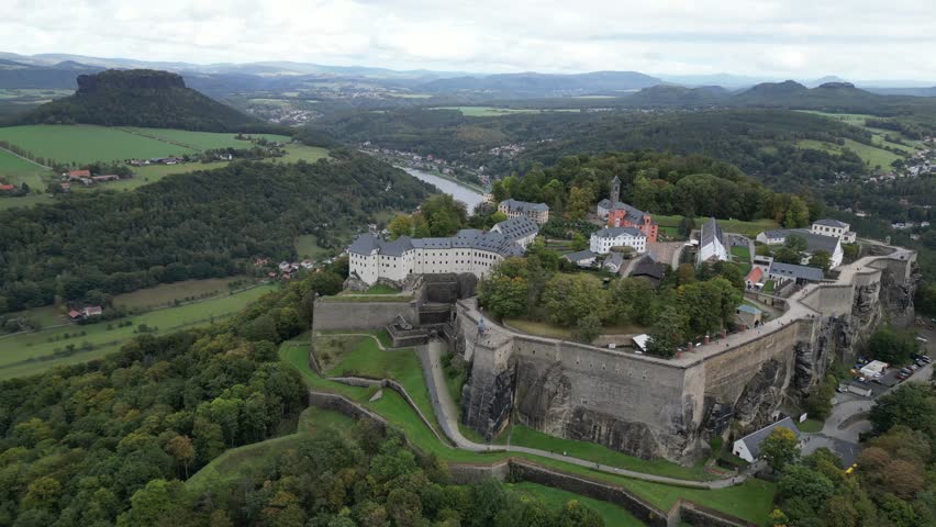An aerial footage of majestic Königstein Fortress, aka the Saxon Bastille, perching on a hilltop and overlooking Lilienstein mesa hill across Elbe River in the green field, south-east of Dresden, in Saxon Switzerland, Germany.

A short aerial video of the medieval Königstein Fortress on a hilltop by a river bend of the Elbe and the beautiful landscape of Saxon Switzerland in Saxony, Germany.

High Stone Walls: Massive, vertical stone fortifications encircle the entire hilltop plateau.
Historical Buildings: Multiple large structures featuring distinct gray tiled roofs sit within the walls.

The Elbe River: A wide, winding blue river curves gracefully through the deep green valley in the background.
Saxon Switzerland Terrain: Dense green forests cover the rolling hills surrounding the fortress plateau.
Distinct Rock Formations: The large, flat-topped mesa hill (Lilienstein table mountain) rises prominently on the horizon.